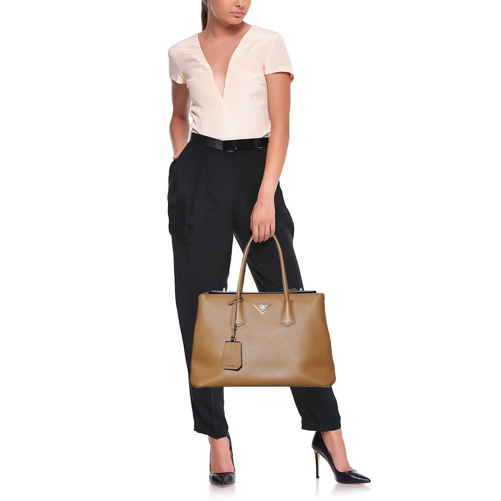 This elegant tote from Prada is crafted from Saffiano leather and is perfect for daily use. The bag features double handles, a leather tag, front logo detail, and protective metal feet at the bottom. The leather-lined interior is spacious enough to