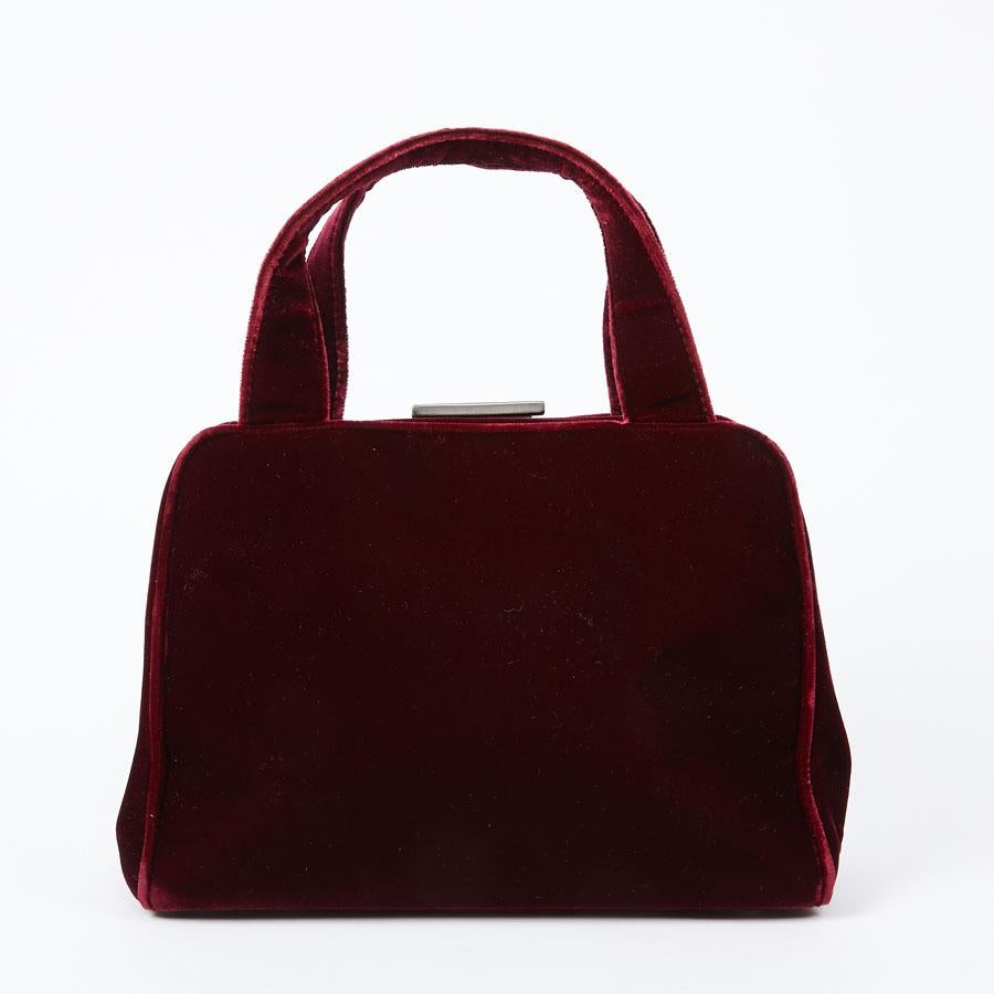 Small Prada red velvet handbag to carry by hand with its double handle. Closing with a rocker button. It is lined in monogram fabric with a zipped pocket. The name 