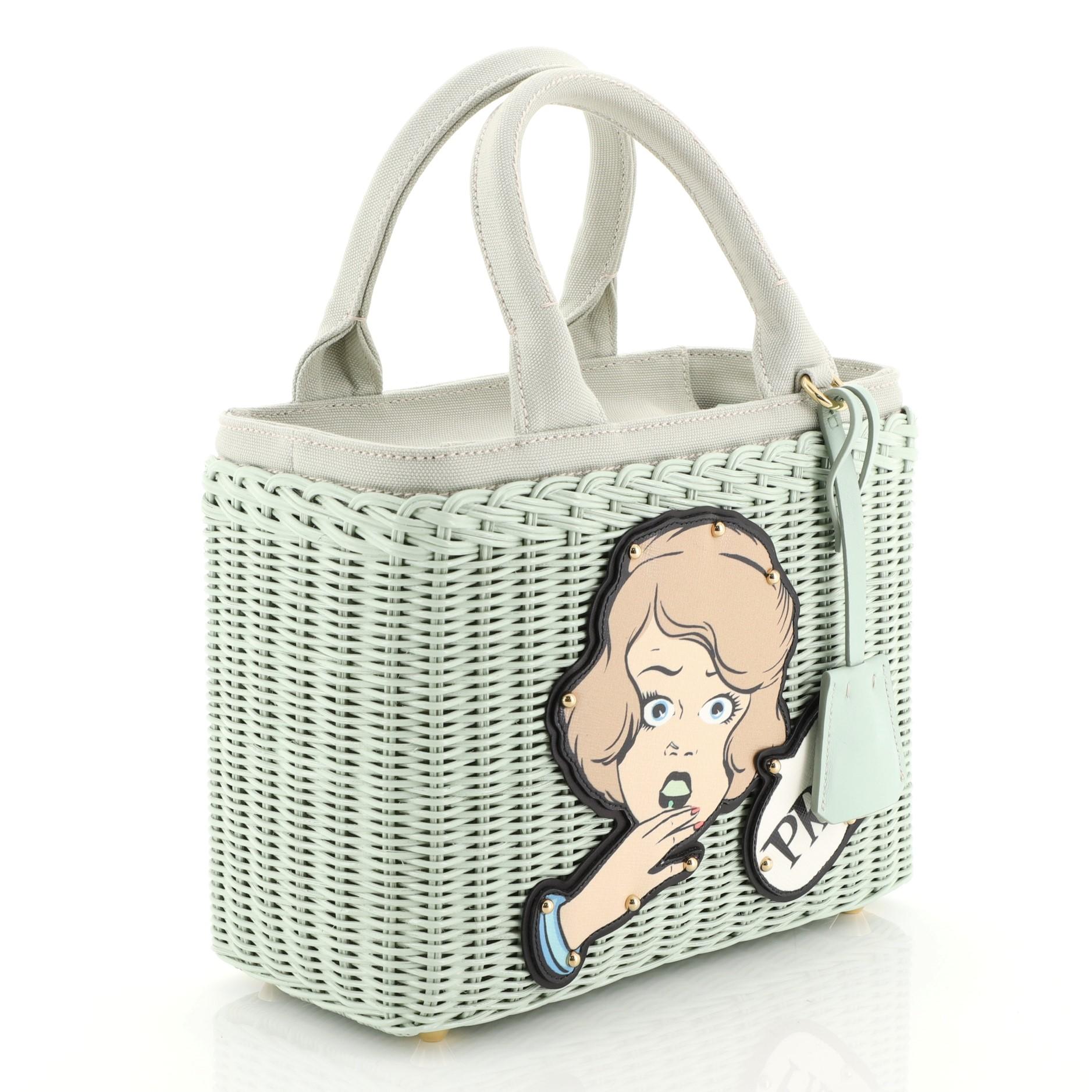 This Prada Comic Basket Bag Wicker with Canapa and Applique Smalll, crafted from green wicker with applique, features dual top handles, protective base studs, and gold-tone hardware. Its zip closure opens to a green fabric interior with zip and slip