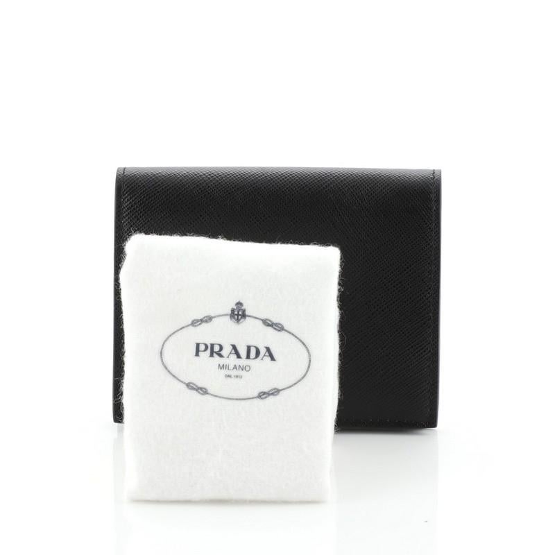 This Prada Compact Monochrome Wallet Saffiano Leather, crafted in black saffiano leather, features raised Prada logo and gold-tone hardware. Its snap button closure opens to a black leather and fabric interior with multiple card slots and zip