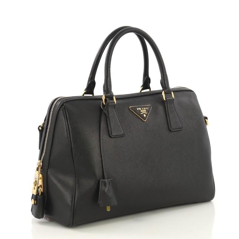 This Prada Convertible Bowler Bag Saffiano Leather Medium, crafted from black saffiano leather, features dual rolled top handles, protective base studs, and gold-tone hardware. Its top zip closure opens to a black fabric interior with zip and slip