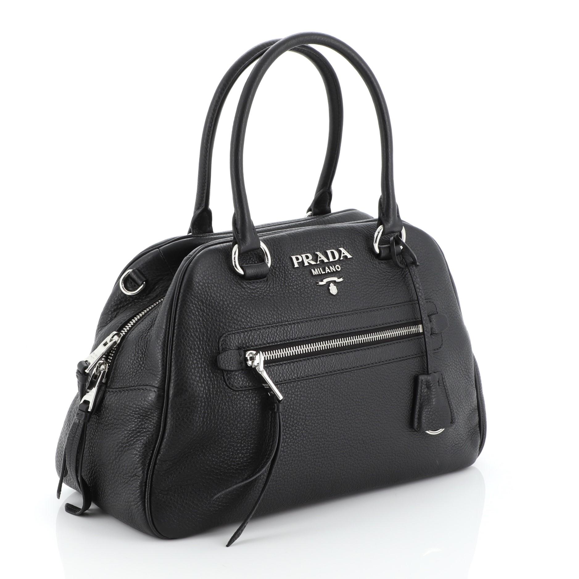 This Prada Convertible Bowling Bag Vitello Daino Medium, crafted in black vitello daino leather, features dual rolled handles, Prada logo at the center, and silver-tone hardware. Its zip closure opens to a black fabric interior with side zip and