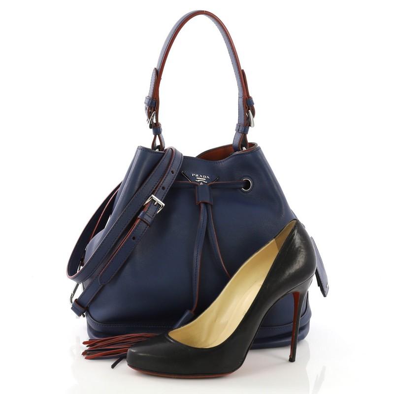 This Prada Convertible Drawstring Bucket Bag Leather Large, crafted in blue leather, features a flat leather handle, drawstring closure with leather tassel accents, and silver-tone hardware. Its drawstring closure opens to a red leather interior