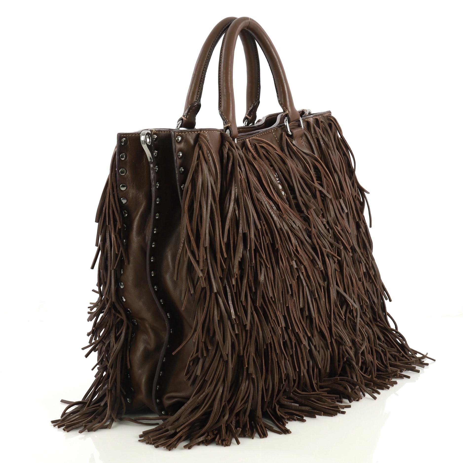 Diese Prada Convertible Fringe Tote Studded Nappa Leather Medium ist aus braunem Leder gefertigt und verfügt über zwei gerollte Griffe mit Ringen, ein umgedrehtes Prada-Logo in der Mitte, Lederfransen, Nietenbesatz und silberfarbene Hardware. Der