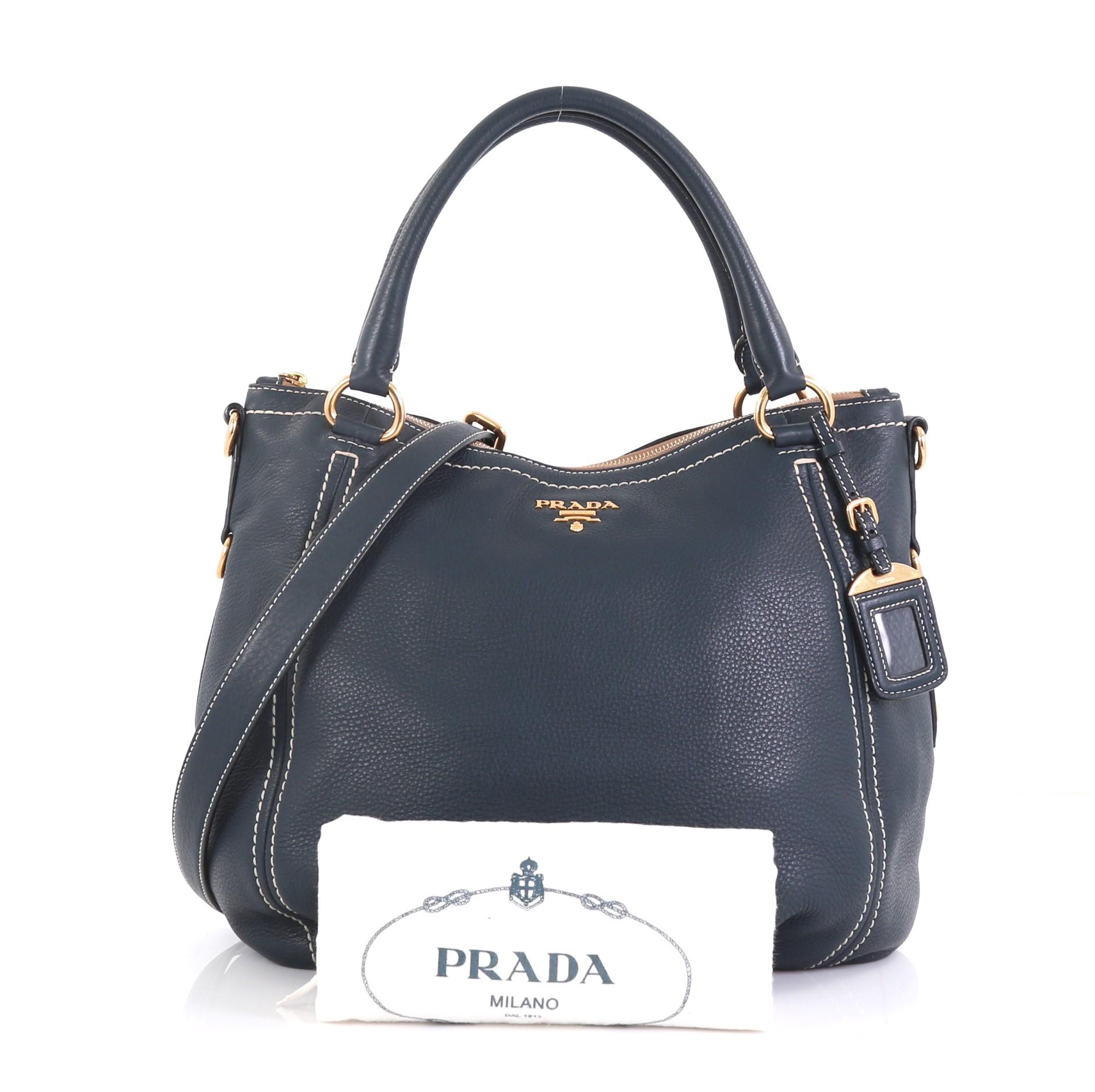 This Prada Convertible Satchel Vitello Daino Large, crafted from navy blue vitello daino leather, features dual rolled handles with rings, Prada logo at its center, and gold-tone hardware accents. Its zip closure opens to a beige fabric and blue