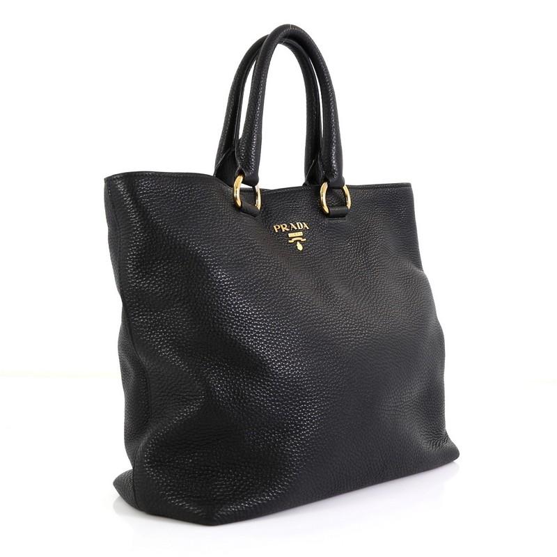This Prada Convertible Shopper Tote Vitello Daino Large, crafted from black vitello daino leather, features dual rolled leather handles, raised Prada logo at the center, and gold-tone hardware. Its magnetic snap closure opens to a black fabric