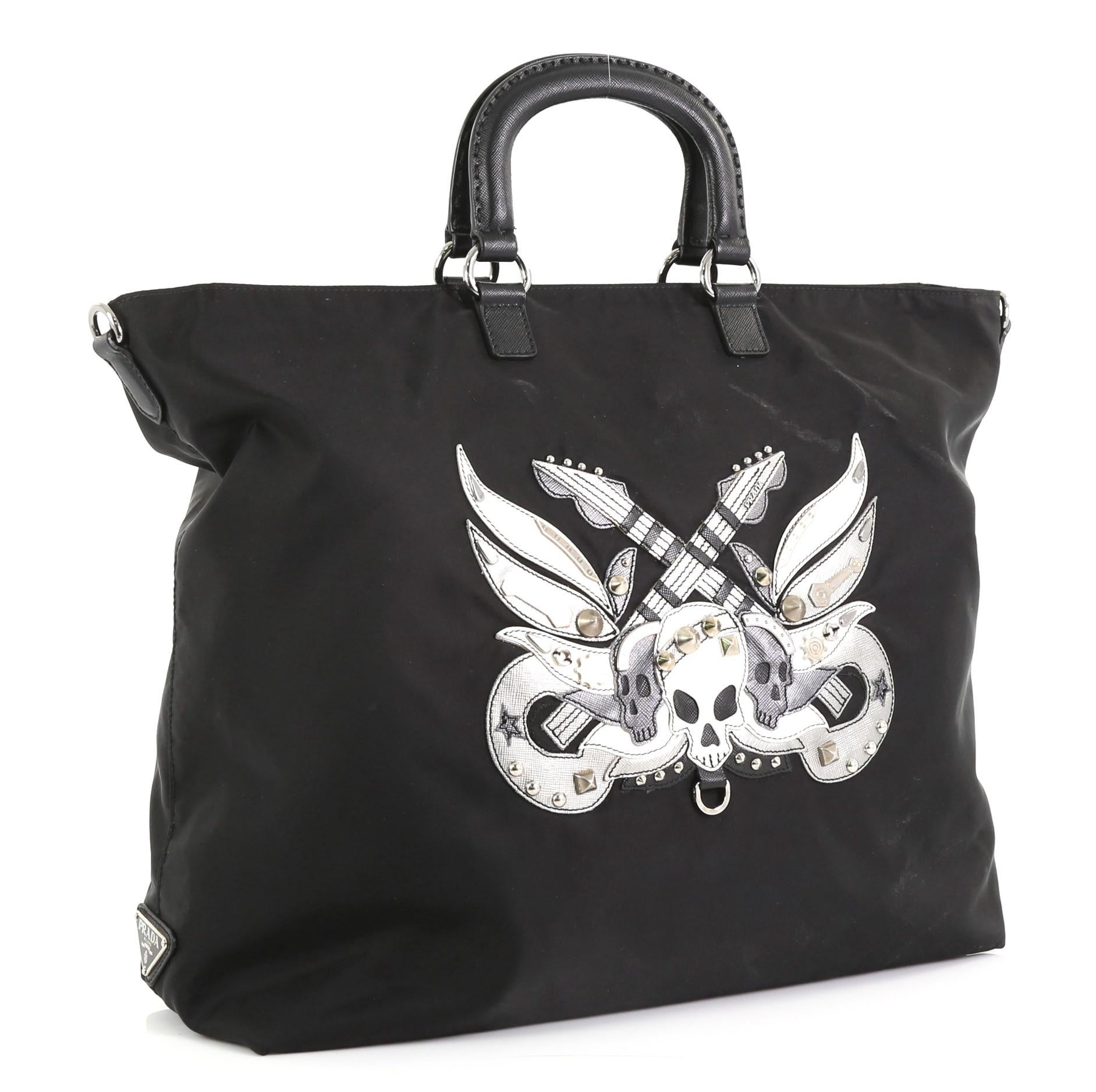 This Prada Convertible Skull Tote Tessuto with Studded Saffiano Leather Large, crafted from black tessuto with studded black saffiano leather, features a stunning skull and guitar leather applique, dual-top leather handles, raised Prada logo and