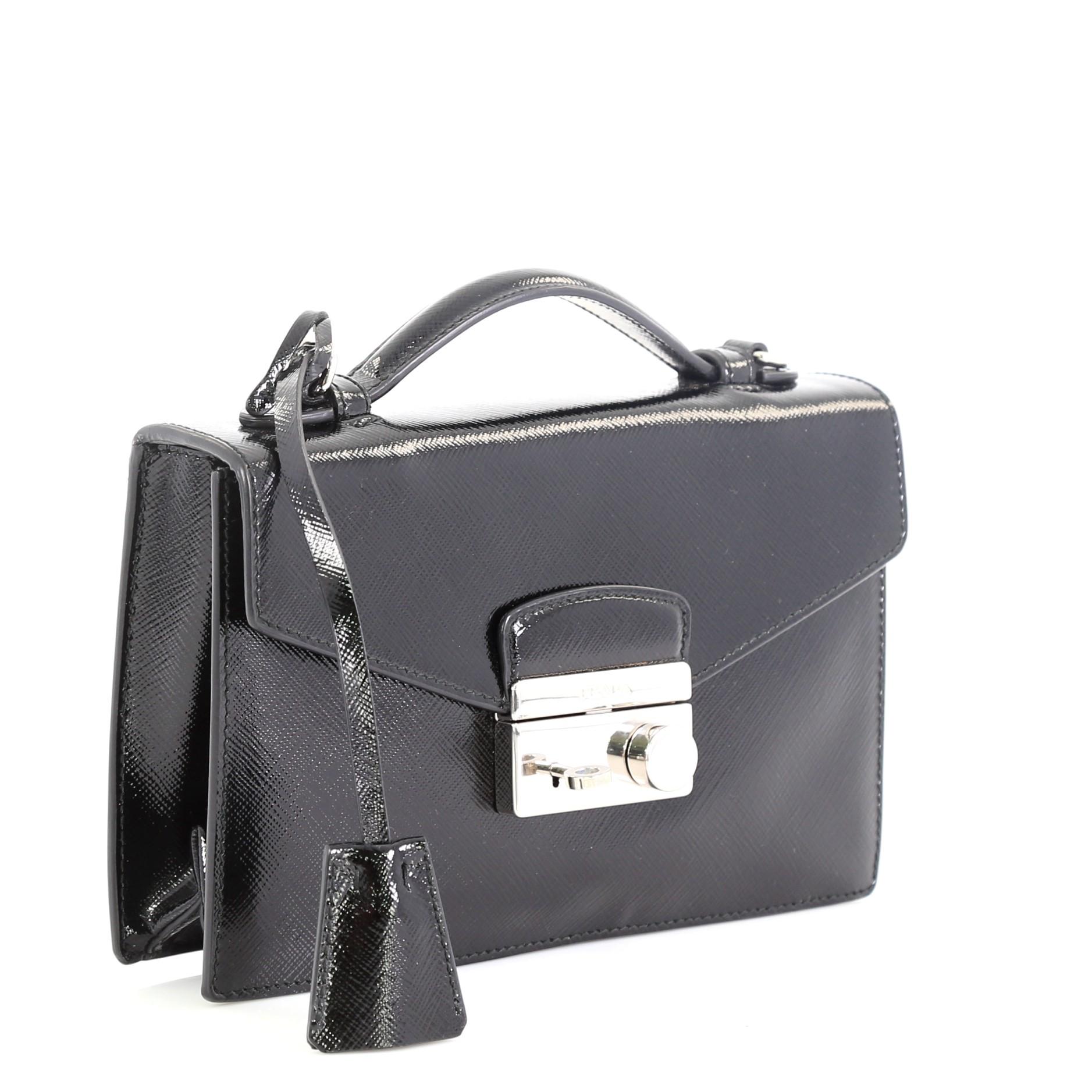This Prada Convertible Sound Bag Vernice Saffiano Leather Small, crafted in black vernice leather, features a short top leather handle, frontal flap with engraved side press closure, long strap and silver-tone hardware. Its press-lock closure opens