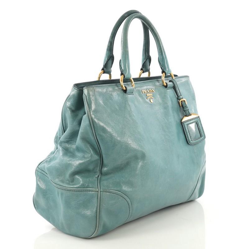 This Prada Convertible Tote Vitello Shine Large, crafted from teal vitello shine leather, features dual rolled handles with rings, raised metal Prada logo at its center, protective base studs, and gold-tone hardware. Its snap button closure opens to