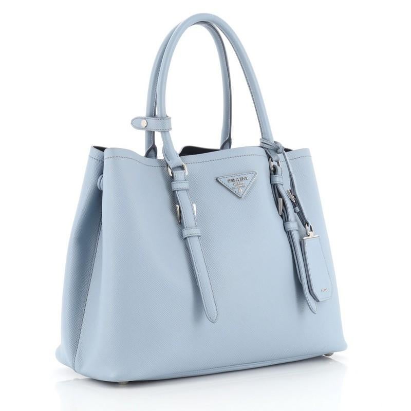 This Prada Covered Strap Cuir Double Tote Saffiano Leather Medium, crafted from blue saffiano leather, features dual rolled handles with extended belt accents, protective base studs, side snap buttons, and silver-tone hardware. It opens to a blue