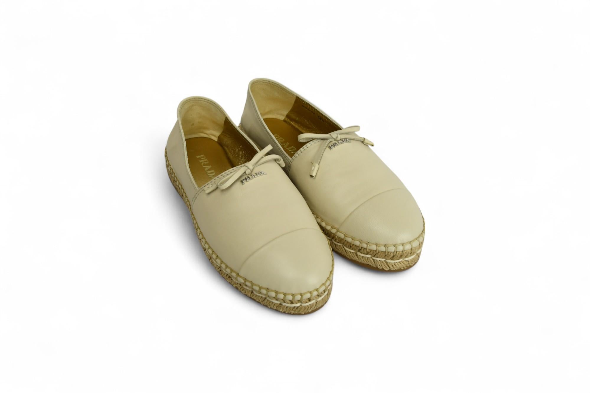These chic Prada espadrilles deliver refined ease with signature Italian craftsmanship. Made in soft cream-toned leather, they feature a rounded cap toe, braided jute midsole, and tonal bow accent with silver Prada logo hardware. The gold metallic
