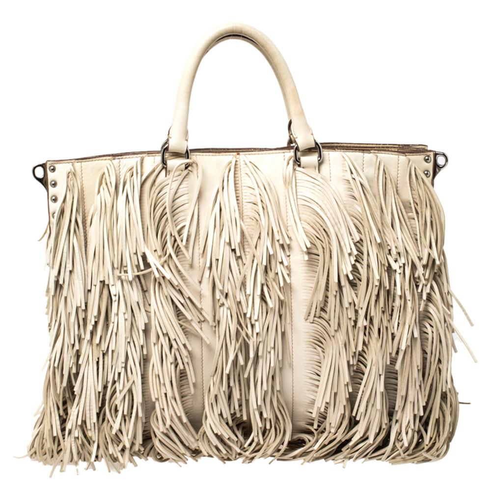 Designed in a large spacious tote size, this Prada bag is high on style as it is high on utility. Crafted in creamish-white leather, this bag is accented with fringe details all through the front and the back of this bag, along with silver-tone stud