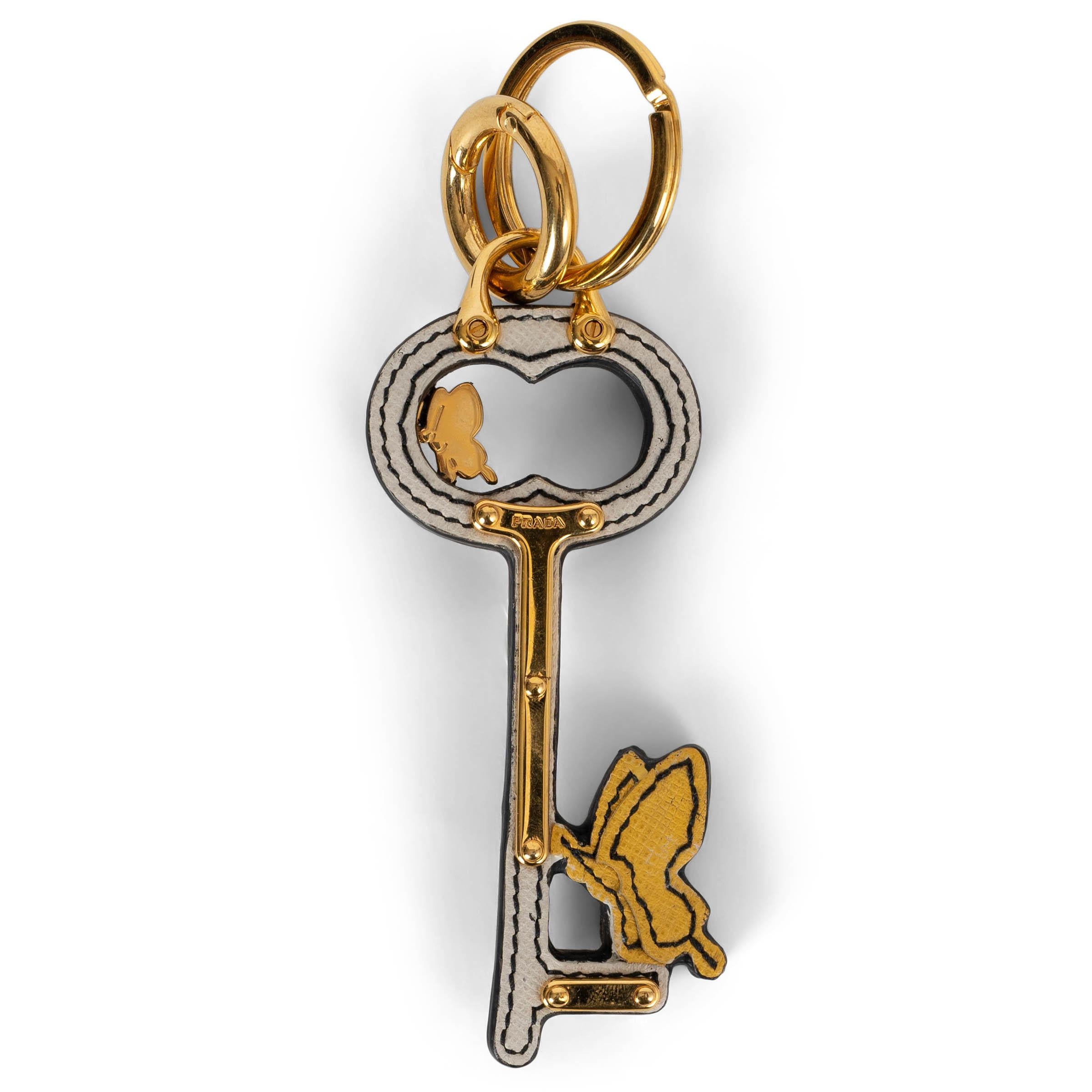 This 100% authentic Prada keyring is crafted in white Saffiano leather shaped like a vintage key, adorned with a yellow butterfly motif and gold-tone metal details. Both whimsical and refined, it adds a playful yet luxurious touch to your keys or