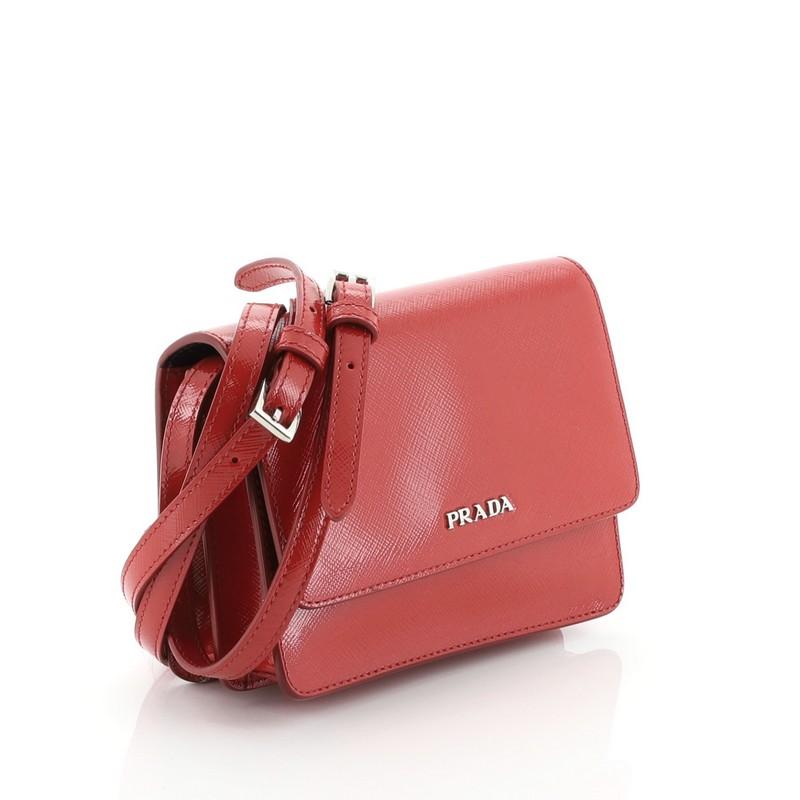 This Prada Crossbody Bag Vernice Saffiano Leather Mini, crafted from red vernice saffiano leather, features an adjustable shoulder strap, raised Prada logo, and silver-tone hardware. Its snap button closure opens to a black leather interior.