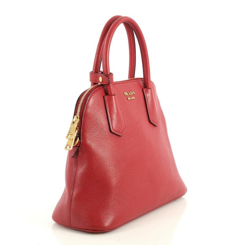 This Prada Cuir Convertible Dome Tote Saffiano Leather Medium, crafted from red saffiano leather, features dual rolled handles, protective base studs, and gold-tone hardware. Its zip closure opens to a red leather interior with zip and slip pockets.