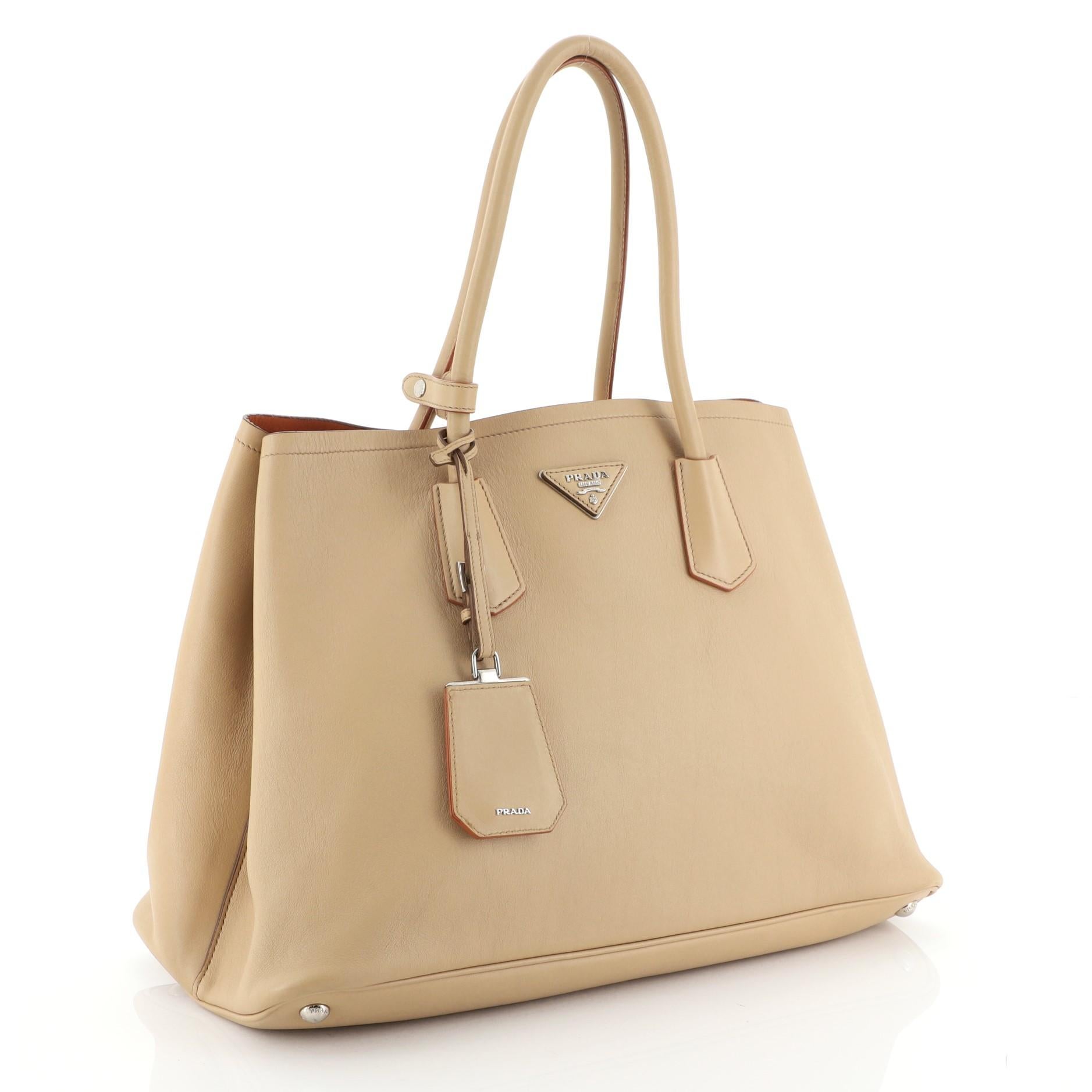 This Prada Cuir Double Tote City Calfskin Large, crafted from neutral calfskin leather, features dual-rolled handles, side snap buttons, protective base studs, Prada's trademark triangle logo at the center and silver-tone hardware accents. It opens