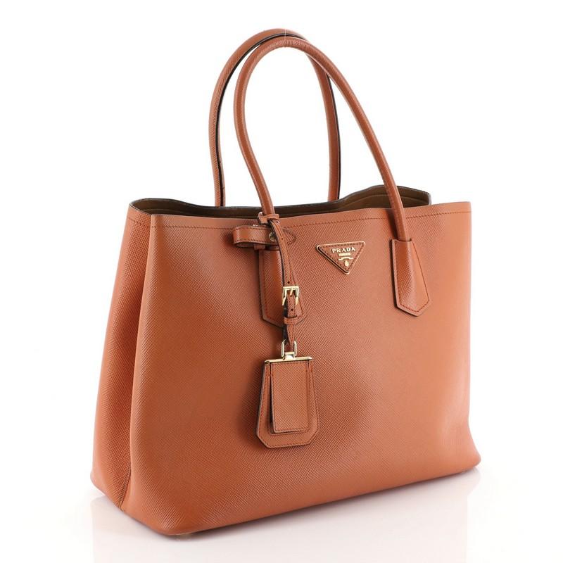 This Prada Cuir Double Tote Saffiano Leather Large, crafted from orange saffiano leather, features dual rolled handles, triangle logo at the center, and gold-tone hardware. It opens to a brown leather interior with middle flap compartment.