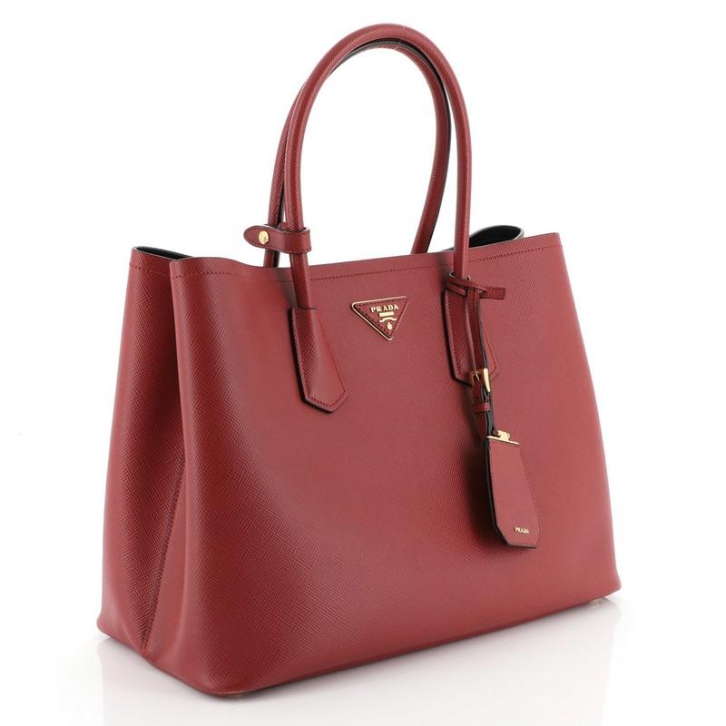 This Prada Cuir Double Tote Saffiano Leather Large, crafted from red saffiano leather, features dual rolled handles, triangle logo at the center, and gold-tone hardware. It opens to a black leather interior with middle flap compartment. 

Estimated