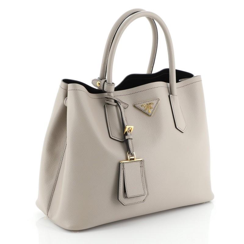 This Prada Cuir Double Tote Saffiano Leather Medium, crafted from neutral saffiano leather, features dual rolled handles, triangle logo at the center, and gold-tone hardware. It opens to a black leather interior with middle flap compartment.