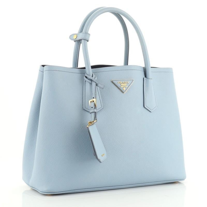 This Prada Cuir Double Tote Saffiano Leather Medium, crafted from blue saffiano leather, features dual rolled handles, triangle logo at the center, and gold-tone hardware. It opens to a blue leather interior with middle flap compartment. 

Estimated