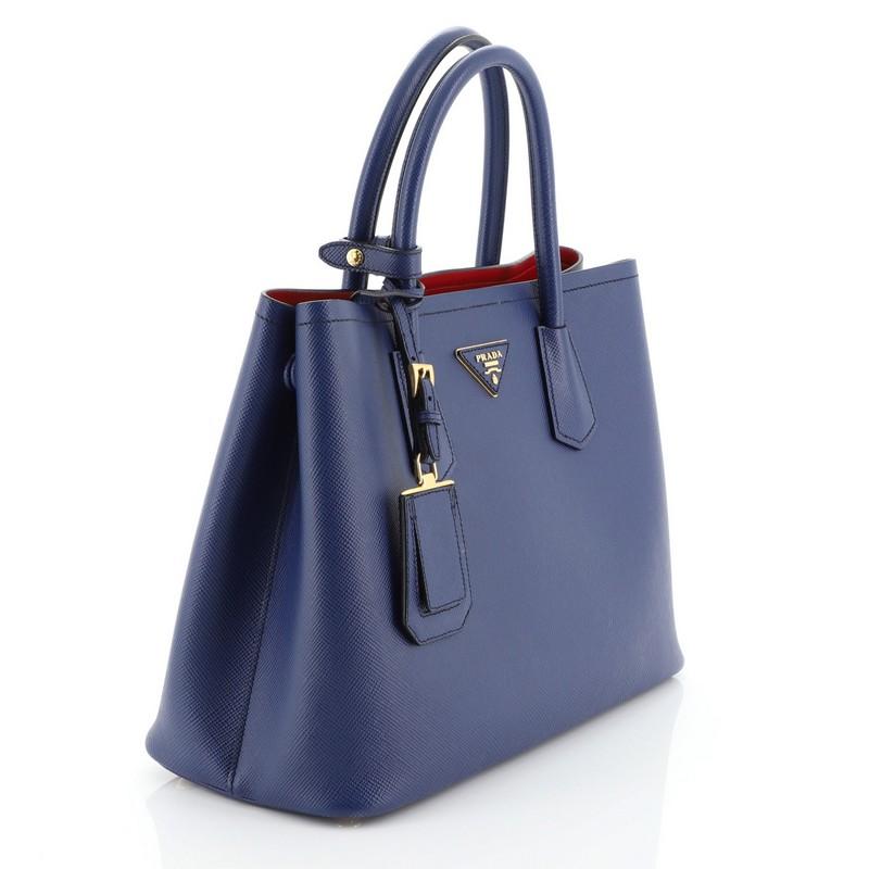 This Prada Cuir Double Tote Saffiano Leather Small, crafted from blue saffiano leather, features dual rolled handles, triangle logo at the center, and gold-tone hardware. It opens to a red leather interior with middle flap compartment. 

Estimated