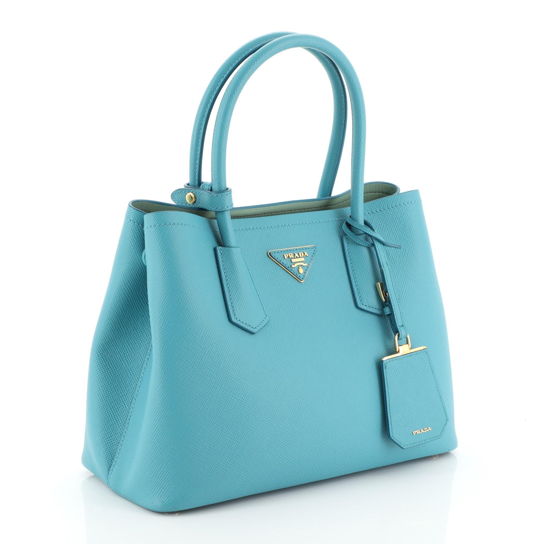 This Prada Cuir Double Tote Saffiano Leather Small, crafted from blue saffiano leather, features dual rolled handles, triangle logo at the center, and gold-tone hardware. It opens to a green leather interior with middle flap compartment. 

Estimated