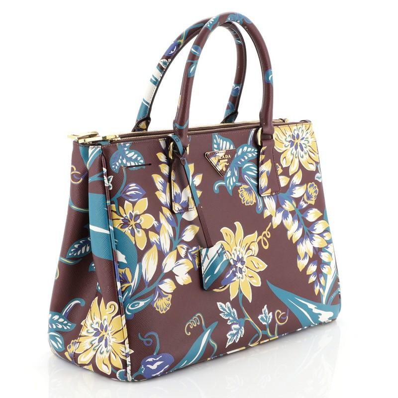 This Prada Double Zip Lux Tote Printed Saffiano Leather Medium, crafted from red printed saffiano leather, features dual rolled handles, raised Prada logo plate, and gold-tone hardware. It opens to a blue leather interior with two zip compartments