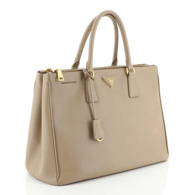 This Prada Double Zip Lux Tote Saffiano Leather Large, crafted from neutral saffiano leather, features dual rolled handles, raised Prada logo plate, and gold-tone hardware. It opens to a neutral fabric interior with two zip compartments on both