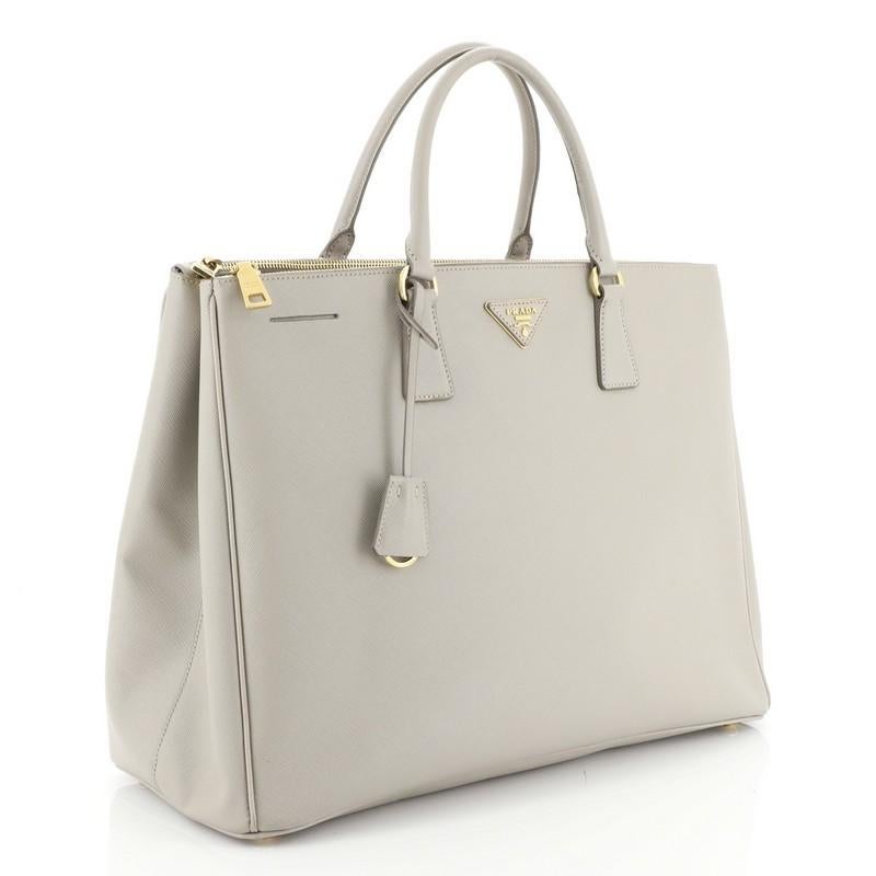 This Prada Double Zip Lux Tote Saffiano Leather Large, crafted from gray saffiano leather, features dual rolled handles, raised Prada logo plate, and gold-tone hardware. It opens to a neutral fabric interior with two zip compartments on both sides