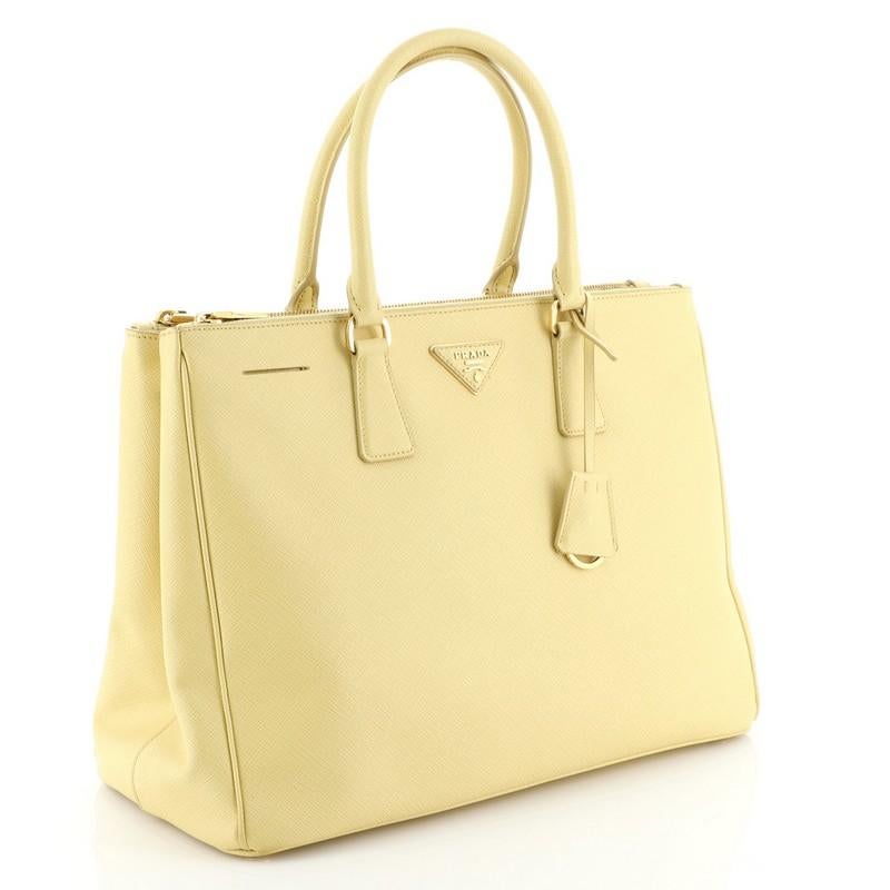 This Prada Double Zip Lux Tote Saffiano Leather Medium, crafted from yellow saffiano leather, features dual rolled handles, raised Prada logo plate, and gold-tone hardware. It opens to a neutral fabric and yellow leather interior with two zip