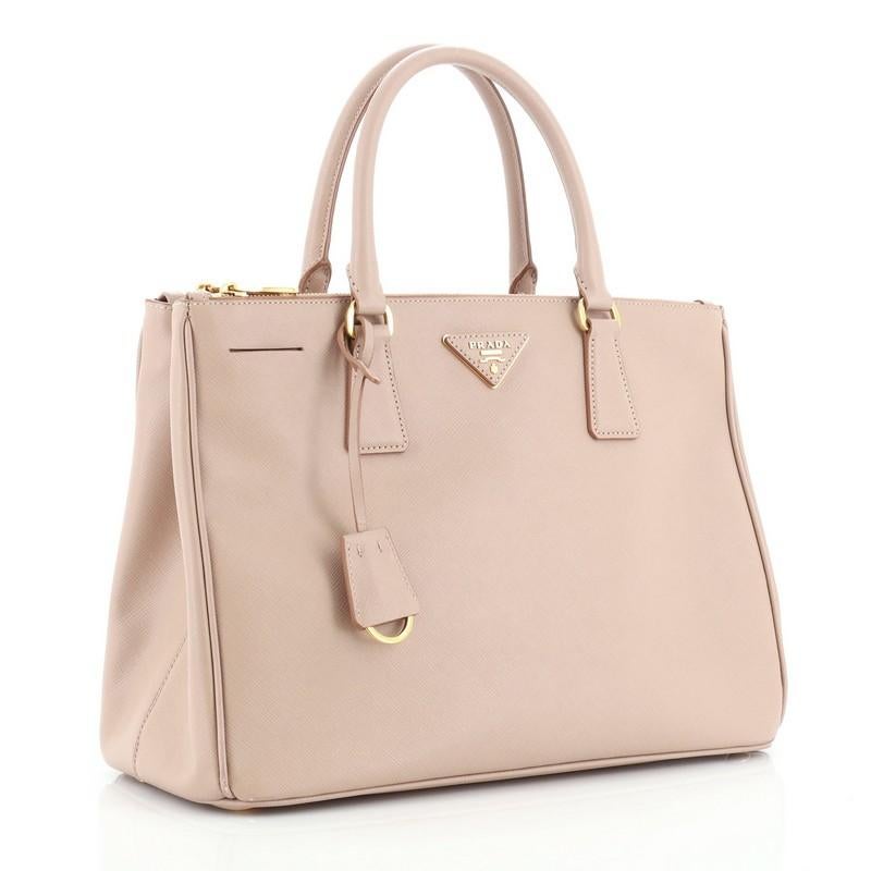 This Prada Double Zip Lux Tote Saffiano Leather Medium, crafted from pink saffiano leather, features dual rolled handles, raised Prada logo plate, and gold-tone hardware. It opens to a pink leather and neutral fabric interior with two zip