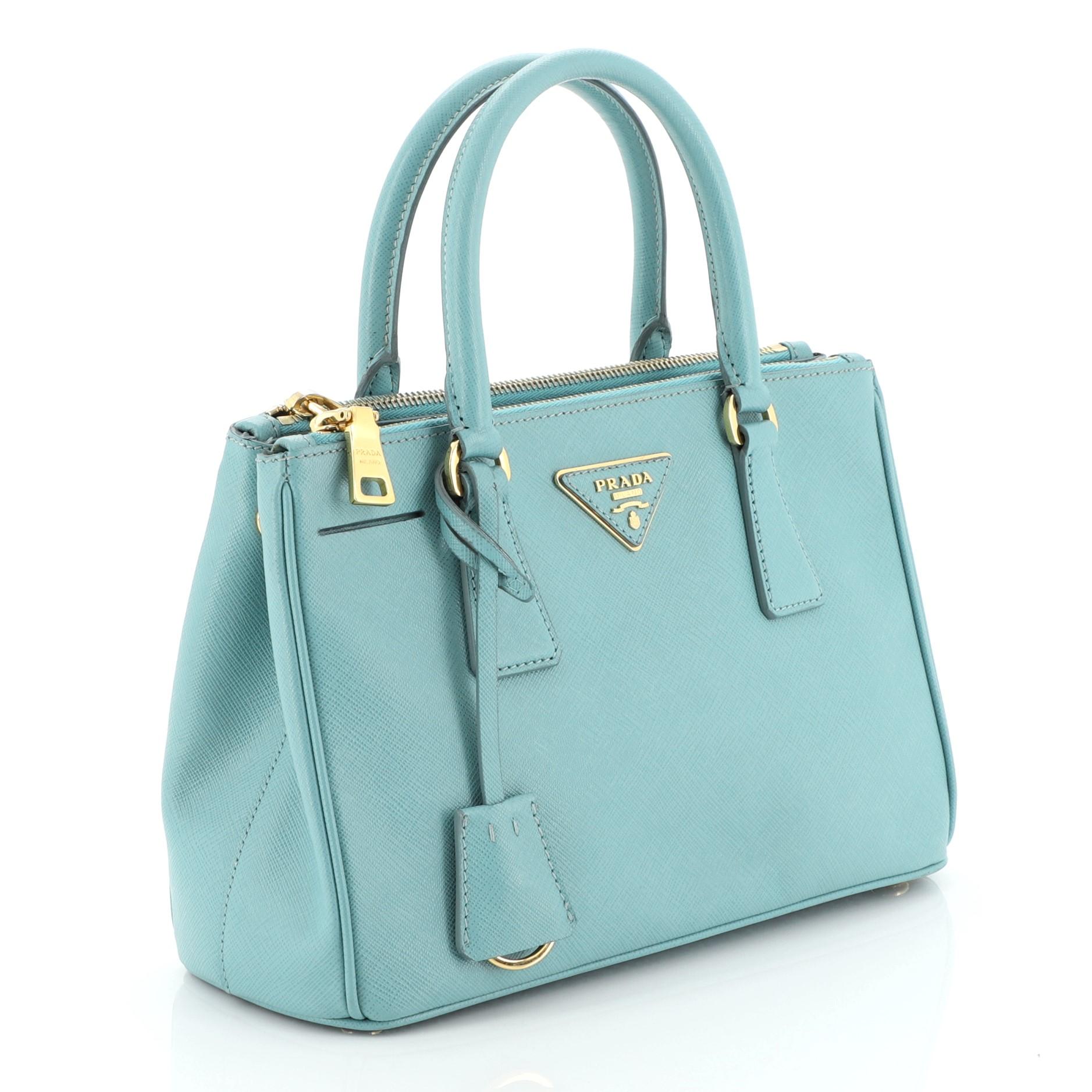 This Prada Double Zip Lux Tote Saffiano Leather Mini, crafted from blue saffiano leather, features dual rolled handles, raised Prada logo plate, and gold-tone hardware. It opens to a blue fabric interior with two zip compartments on both sides and
