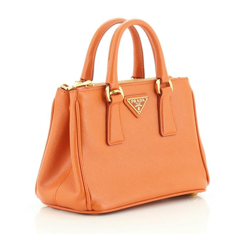 This Prada Double Zip Lux Tote Saffiano Leather Nano, crafted from orange saffiano leather, features dual rolled handles, raised Prada logo plate, and gold-tone hardware. It opens to an orange fabric interior with two zip compartments on both sides