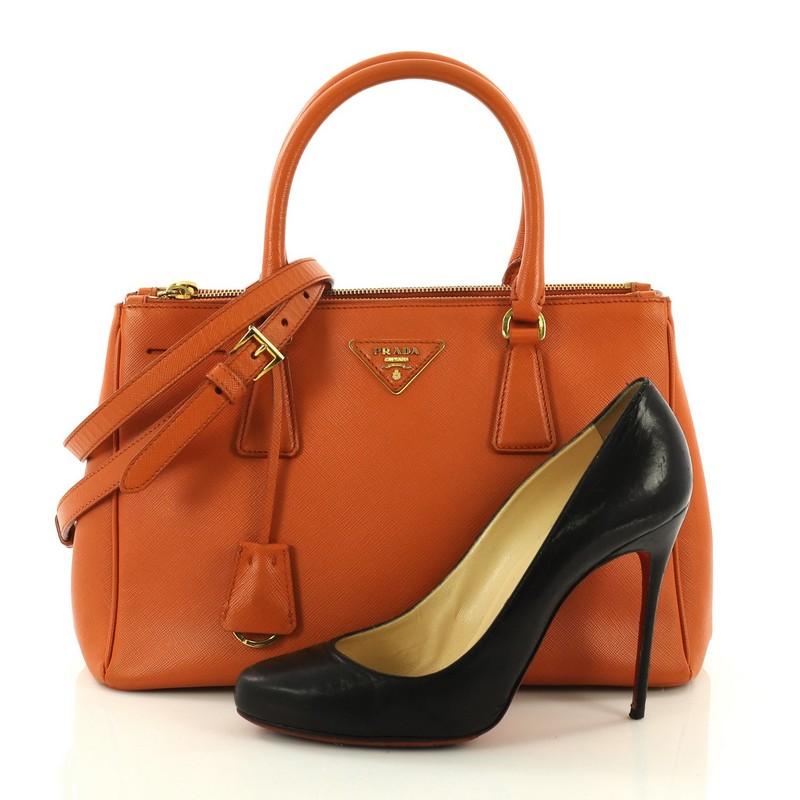 This Prada Double Zip Lux Tote Saffiano Leather Small, crafted from orange saffiano leather, features dual rolled handles, raised Prada logo plate, and gold-tone hardware. It opens to an orange fabric interior with two zip compartments on both sides