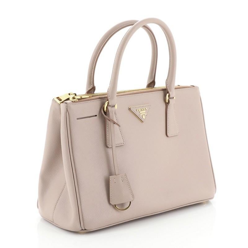 This Prada Double Zip Lux Tote Saffiano Leather Small, crafted from pink saffiano leather, features dual rolled handles, raised Prada logo plate, and gold-tone hardware. It opens to a pink leather and neutral fabric interior with two zip