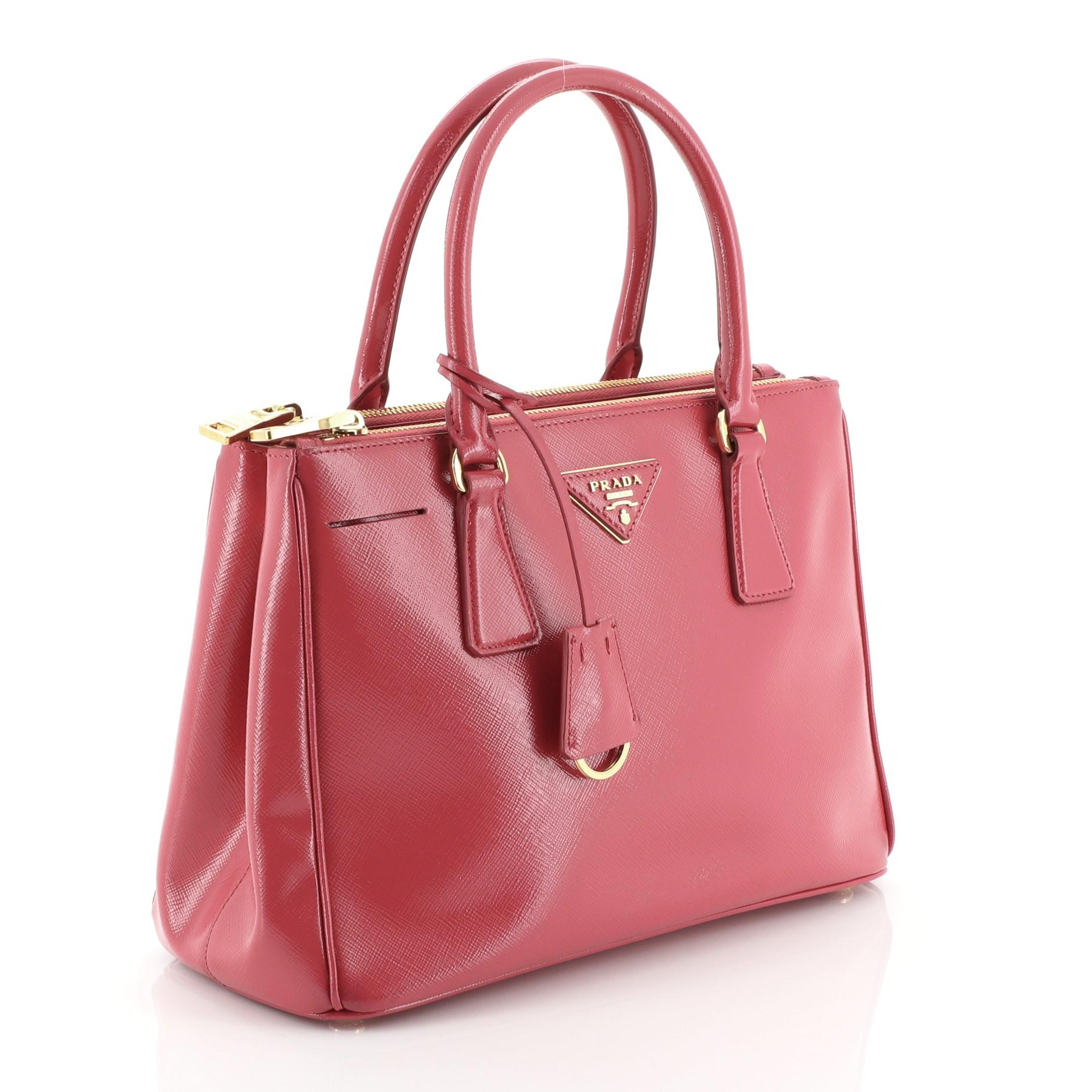 This Prada Double Zip Lux Tote Vernice Saffiano Leather Mini, crafted from pink vernice saffiano leather, features dual rolled handles, raised Prada logo plate, and gold-tone hardware. It opens to a pink fabric interior with two zip compartments on