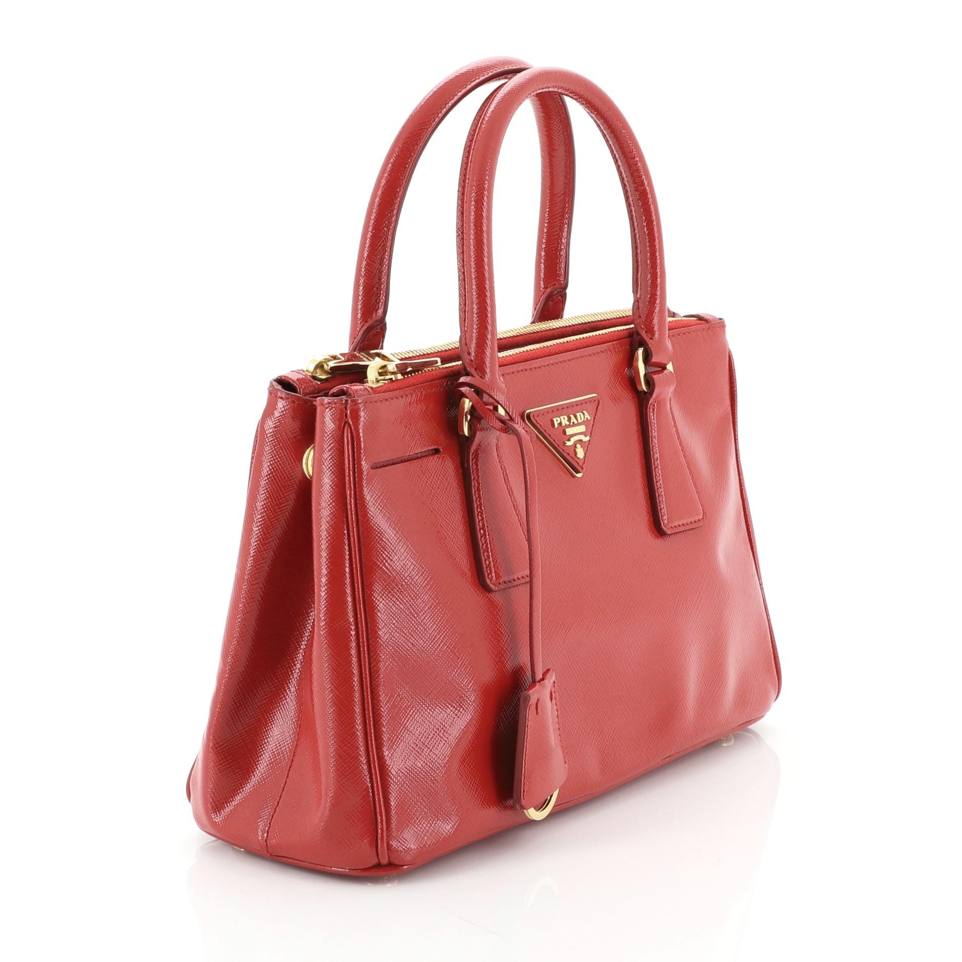 This Prada Double Zip Lux Tote Vernice Saffiano Leather Mini, crafted from red vernice saffiano leather, features dual rolled handles, raised Prada logo plate, and gold-tone hardware. It opens to a red fabric and leather interior with two zip