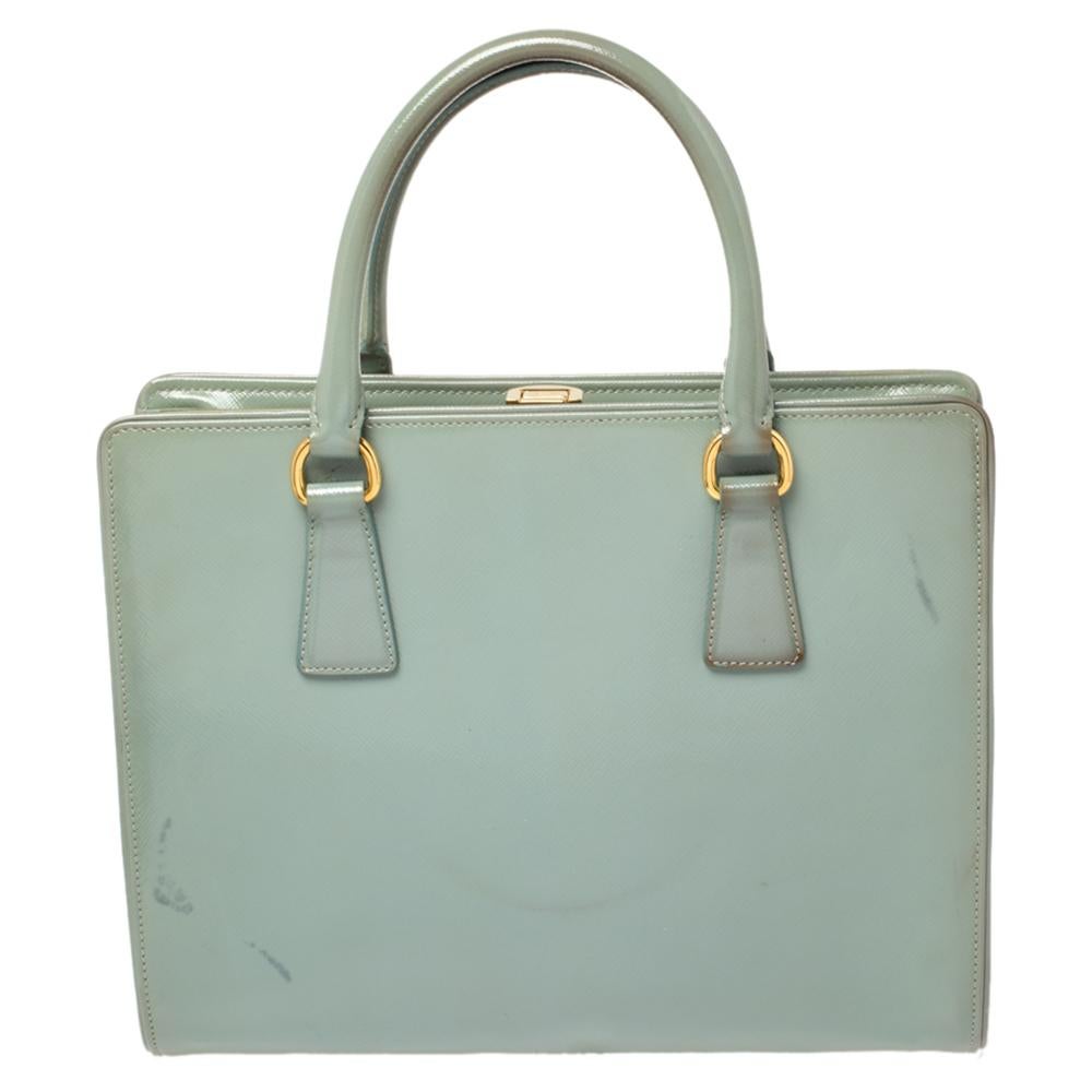 Fashioned in a structured silhouette, this Prada tote is a charming piece that will go well with all your ensembles. It features a dusty blue patent leather body with a top frame secured with a flip-lock closure. Fitted with two sleek shoulder
