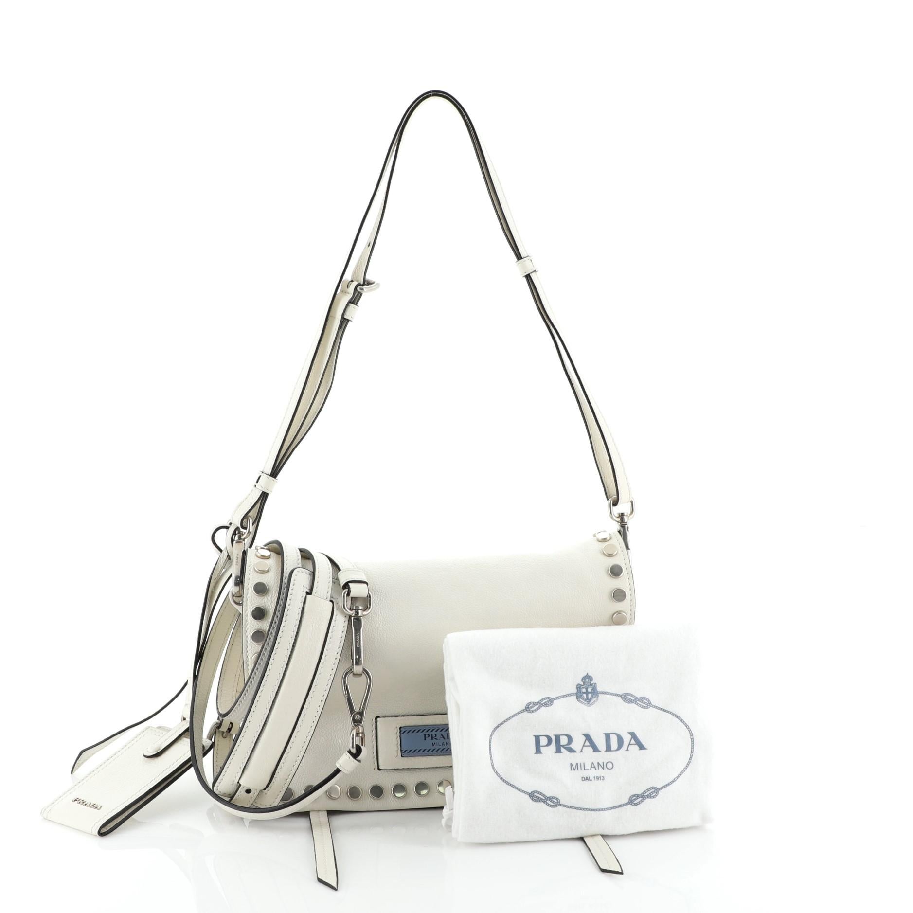 This Prada Etiquette Flap Bag Studded Glace Calfskin Small, crafted from white glace calfskin leather with disc studs, features a detachable shoulder strap, two side zip compartments, and silver-tone hardware. It opens to a blue suede interior