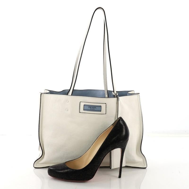This Prada Etiquette Tote Glace Calf Medium, crafted from white glace calf leather, features dual slim leather handles, Prada logo at the front, and silver-tone hardware. It opens to a blue suede interior with a center zip compartment and side slip