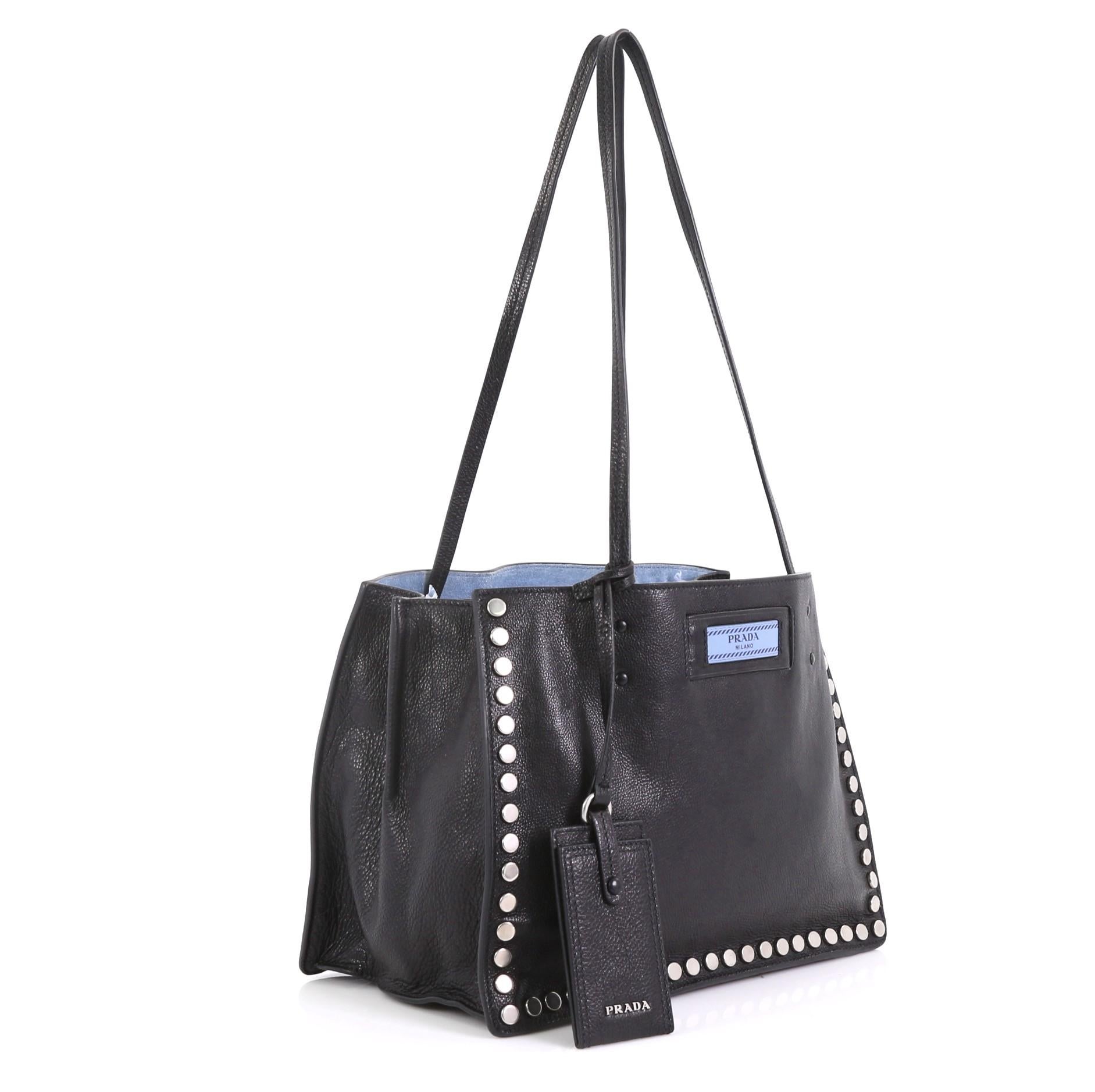 This Prada Etiquette Tote Studded Glace Calfskin Small, crafted from black studded glace calfskin leather, features dual slim leather handles, Prada logo at the front and silver-tone hardware. Its wide open top showcases a blue suede interior with a