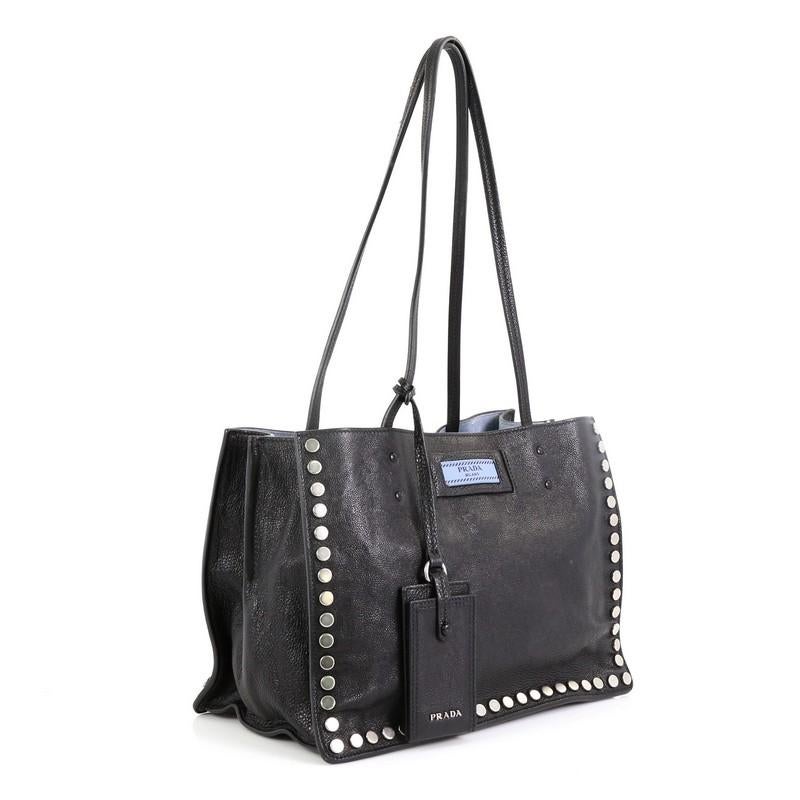 This Prada Etiquette Tote Studded Glace Calfskin Small, crafted from black studded glace calfskin leather, features dual slim leather handles, Prada logo at the front and aged silver-tone hardware. Its wide open top showcases a blue suede interior