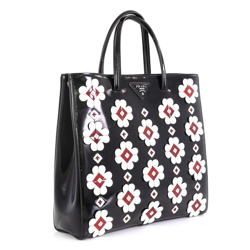 This Prada Flowers Convertible Open Tote Spazzolato Leather Medium, crafted from black leather, features a white flower applique on both sides, dual-rolled top handles, and silver-tone hardware. Its snap button closure opens to a black fabric