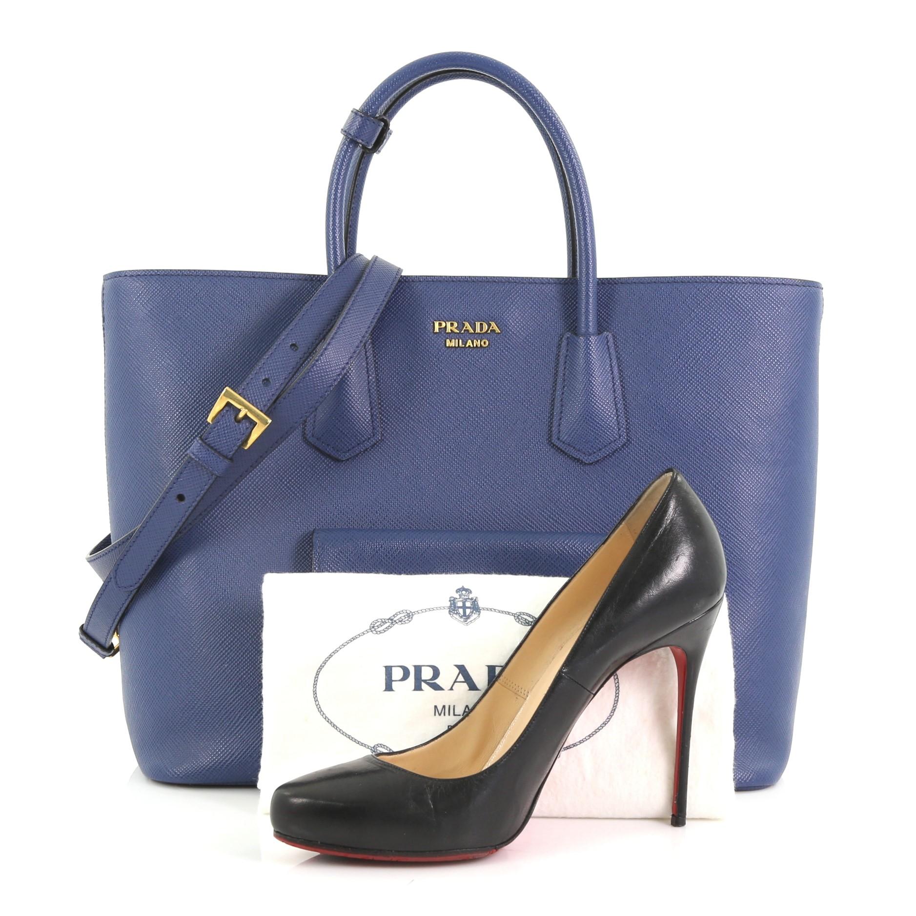 This Prada Front Pocket Convertible Tote Saffiano Leather, crafted from blue saffiano leather, features dual rolled top handles, exterior front snap pockets, and gold-tone hardware. Its snap button closure opens to a blue leather interior with side