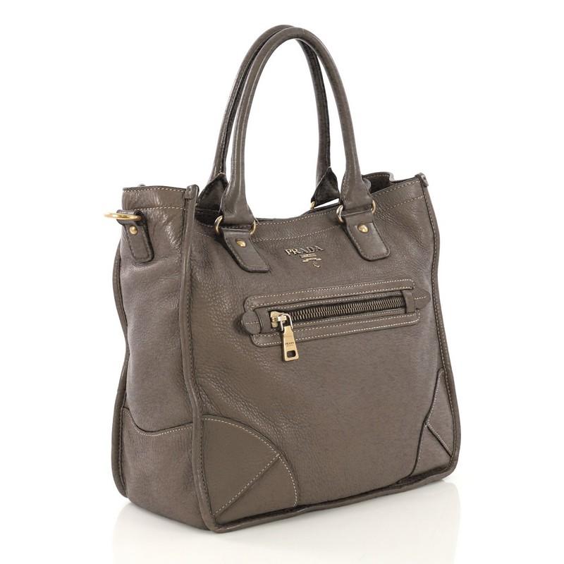 This Prada Front Pocket Convertible Tote Vitello Daino Medium, crafted from gray vitello daino leather, features dual rolled leather handles, exterior zip pockets, signature Prada logo at the center, protective base studs, and aged gold-tone