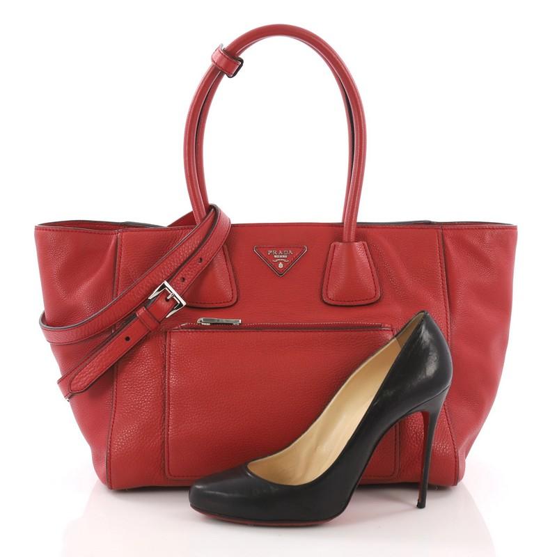 This Prada Front Pocket Wing Convertible Tote Vitello Daino, crafted in red vitello daino leather, features dual rolled leather handles, exterior front zip pocket, and silver-tone hardware. Its snap button closure opens to a black fabric interior