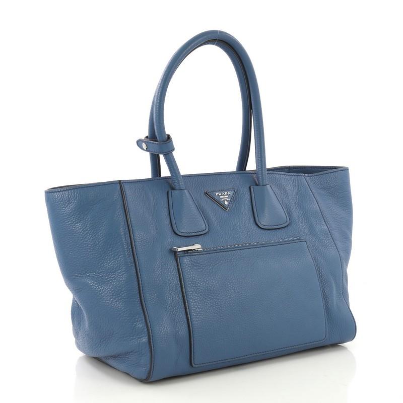 This Prada Front Pocket Wing Convertible Tote Vitello Daino, crafted in blue vitello daino leather, features dual rolled leather handles, exterior front zip pocket, and silver-tone hardware. Its snap button closure opens to a black fabric interior