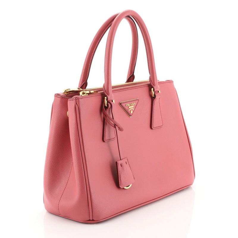 This Prada Galleria Double Zip Tote Saffiano Leather Medium, crafted from pink saffiano leather, features side snap buttons, raised Prada logo plate, dual rolled leather handles, and gold-tone hardware. It opens to a pink fabric interior with two