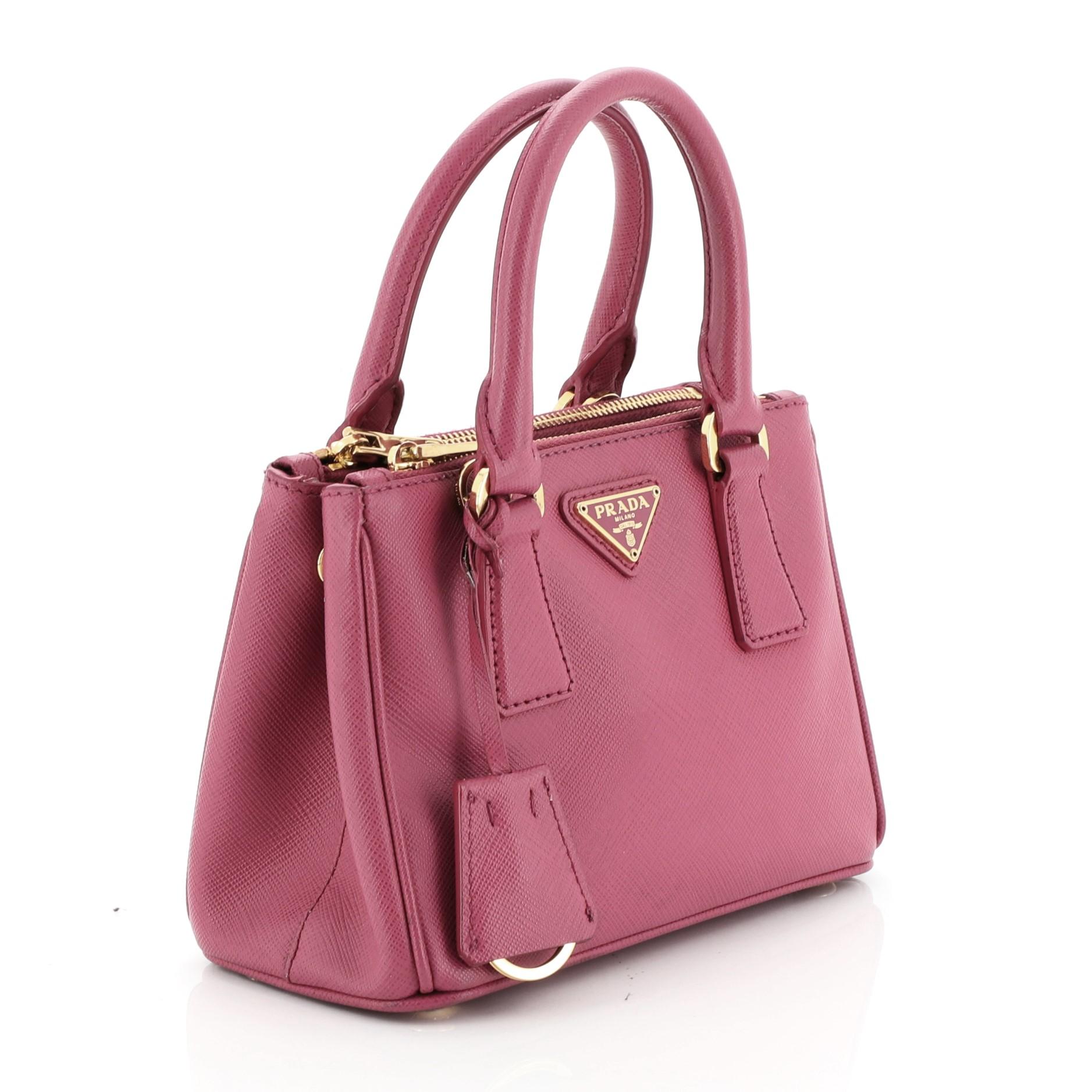 This Prada Galleria Double Zip Tote Saffiano Leather Mini, crafted from pink saffiano leather, features side snap buttons, raised Prada logo plate, dual rolled leather handles, and gold-tone hardware. It opens to a pink fabric interior with two zip