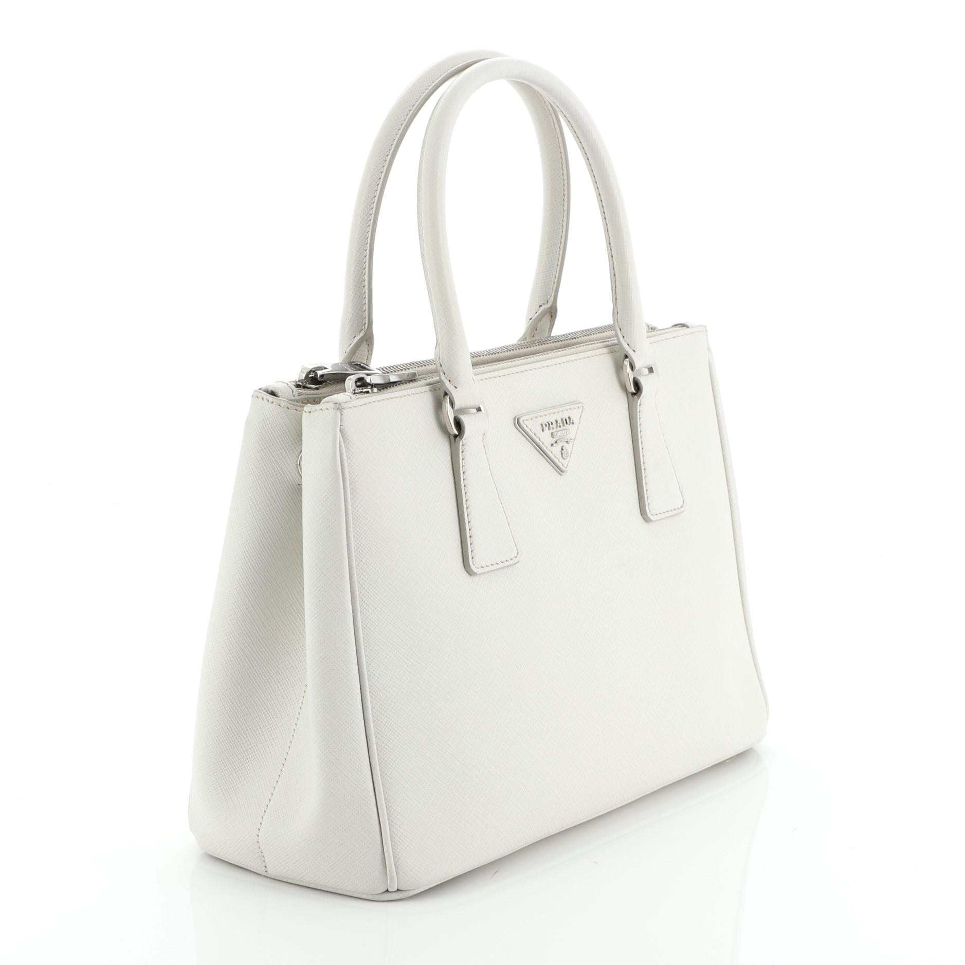 This Prada Galleria Double Zip Tote Saffiano Leather Small, crafted from white saffiano leather, features side snap buttons, raised Prada logo plate, dual rolled leather handles, and silver-tone hardware. It opens to a neutral fabric interior with